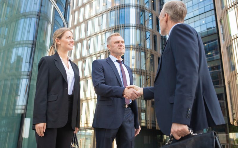 Confident business partners meeting in city, shaking hands near office building. Low angle shot. Cooperation and partnership concept