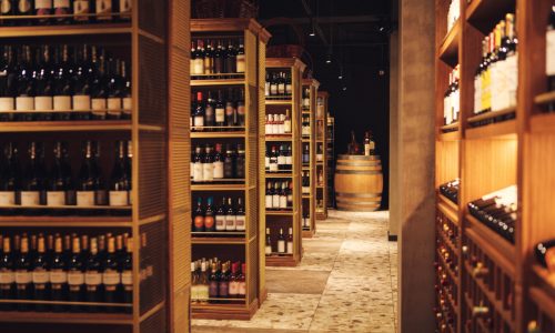 Interior photo of long corridor with wooden shelves fulled bottles of elite wine in cellar of alcohol store. Bottles of various wines lie beautifully on the counter. Big assortment of alcoholic beverages in winery.