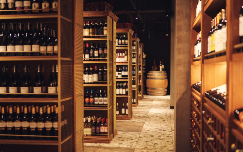 Interior photo of long corridor with wooden shelves fulled bottles of elite wine in cellar of alcohol store. Bottles of various wines lie beautifully on the counter. Big assortment of alcoholic beverages in winery.